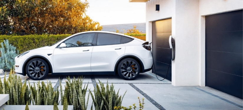 White Tesla charging in the driveway of a modern white house with two black garage doors.